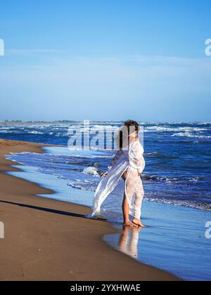 bella donna mediterranea dai capelli ricci incinta posa sulla spiaggia Foto Stock