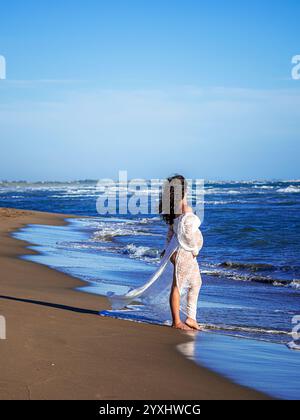 bella donna mediterranea dai capelli ricci incinta posa sulla spiaggia Foto Stock