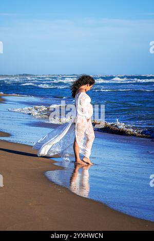 bella donna mediterranea dai capelli ricci incinta posa sulla spiaggia Foto Stock