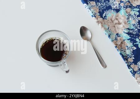 Vista dall'alto verso il basso della tazza da caffè in vetro trasparente e del cucchiaio argentato sul tavolo bianco con tovagliolo in tessuto blu nell'angolo superiore destro. Bibita calda, caffeina. Foto Stock