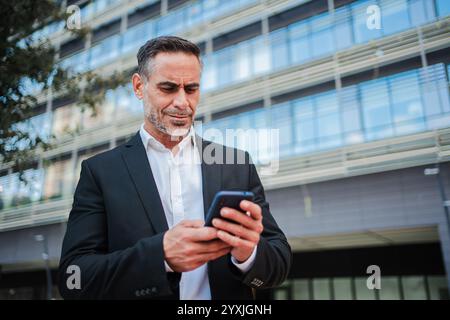 Uomo d'affari maturo e fiducioso in completo stato di guardia all'aperto, che usa un cellulare per navigare online. Uomo d'affari concentrato sull'utilizzo dello smartphone per la comunicazione, il controllo delle e-mail o la gestione delle attività lavorative. Foto di alta qualità Foto Stock