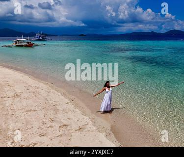 Una splendida giovane donna in un abito bianco che scorre si erge graziosamente su sabbia bianca, con acqua turchese e barche ormeggiate sullo sfondo, evocando un se Foto Stock