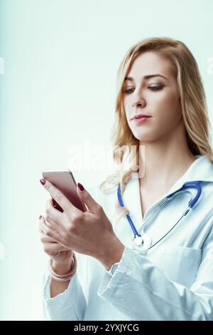 Una giovane dottoressa che indossa un camice da laboratorio e uno stetoscopio è impegnata a leggere notizie mediche sul suo smartphone mentre naviga negli ambienti frenetici Foto Stock