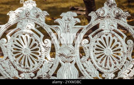 Primo piano della panca da giardino in stile vittoriano in ghisa dipinta di bianco nel parco cittadino. Luce solare sulla superficie dello schienale di un banco in ghisa antica decorata vuota in Foto Stock