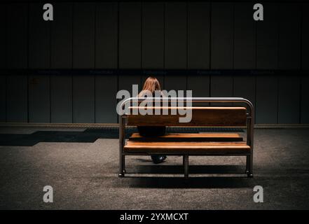 Una donna solitaria con lunghi capelli marrone-rossastro siede su una panchina di legno con una struttura in metallo. La panca è situata in una stazione della metropolitana poco illuminata con wal grigio Foto Stock