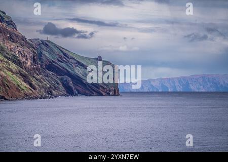 Una suggestiva vista di una scogliera costiera coperta di vegetazione, che contrasta con le calme acque oceaniche e la lontana catena montuosa sotto un cielo nuvoloso, evok Foto Stock