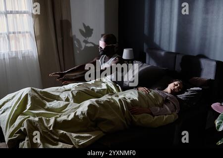 Colpo ad angolo alto di un uomo afroamericano che soffre di sonnambulismo alzandosi dal letto mentre la moglie dorme Foto Stock