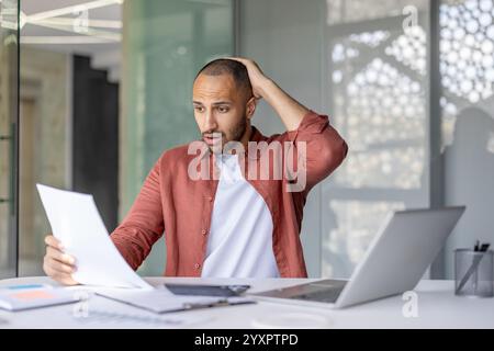 Un uomo che indossa una camicia casual sembra sorpreso mentre esamina i documenti in un ufficio elegante, riflettendo uno scenario di scoperta inaspettato o correlato al lavoro. Foto Stock
