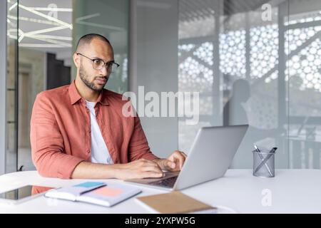 Un individuo attento che indossa gli occhiali lavora diligentemente su un laptop in ufficio, trasmettendo professionalità, produttività e concentrazione, con forniture per ufficio e interni curati Foto Stock