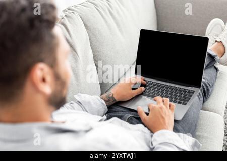 Concetto di pubblicità. Giovane ragazzo che utilizza un moderno computer portatile con schermo vuoto, seduto sul divano, mockup, sopra le spalle Foto Stock