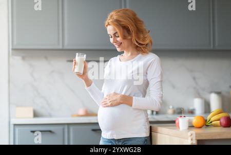 La mamma che si aspetta si gode un bicchiere di latte nella cucina moderna durante il giorno Foto Stock