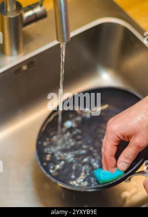 lavare a mano una padella nera in un lavandino in acciaio inox con una spugna blu. Foto di alta qualità Foto Stock