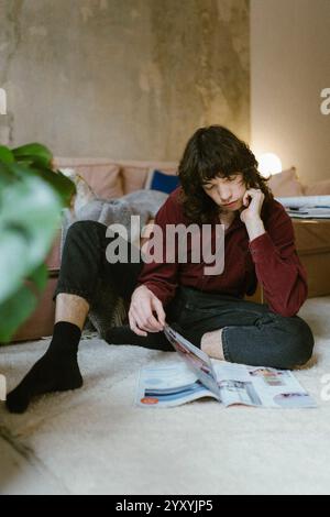 Giovane che legge una rivista mentre seduto sul pavimento a casa Foto Stock