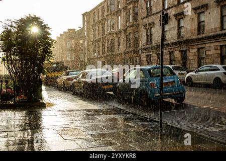 Una strada di città con auto parcheggiate sotto la pioggia battente con il sole che splende. La scena è ambientata in un ambiente urbano, caratterizzato da una fila di veicoli Foto Stock