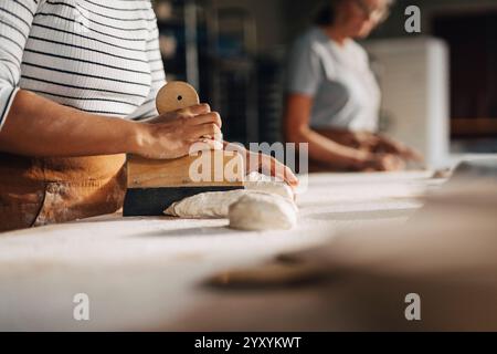 Sezione centrale della panettiera femminile che taglia l'impasto con il taglierino mentre lavora in un panificio Foto Stock