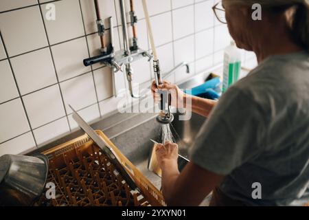 Vista ad angolo alto della panificatrice senior che spruzza acqua sui piatti mentre lavora in cucina commerciale Foto Stock