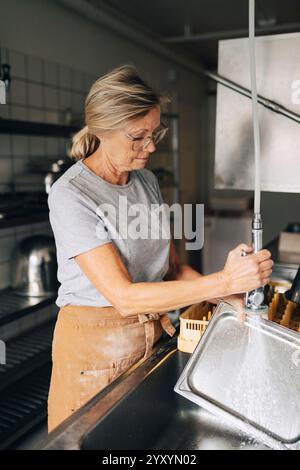 panettiera senior che spruzza acqua sul vassoio mentre lavora in cucina commerciale Foto Stock