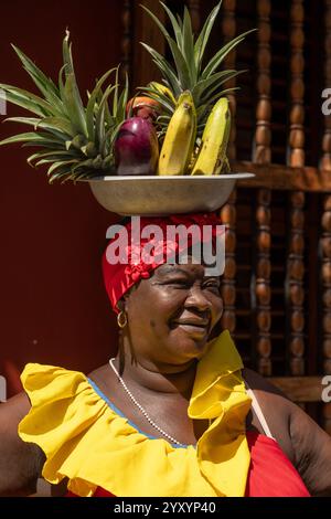 Donna colombiana sorridente che indossa abiti tradizionali e frutta tropicale bilanciata sulla testa davanti alla parete rossa. Foto Stock