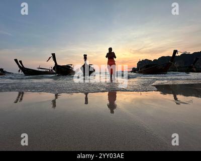 Un uomo sta su una spiaggia con barche al tramonto sullo sfondo Foto Stock