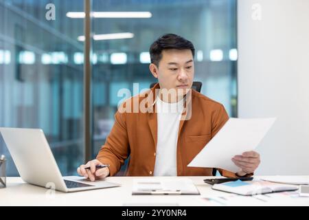 Uomo asiatico uomo d'affari maturo in un ufficio che esamina i documenti mentre lavora su un notebook, rappresentando produttività, professionalità e concentrazione in un ambiente aziendale. Foto Stock