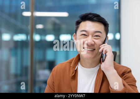 Uomo asiatico che parla al telefono con un sorriso, seduto in un luminoso ambiente d'ufficio. Mostrare fiducia e professionalità, mostrare felicità e buon umore durante la comunicazione Foto Stock