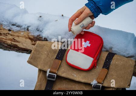 Primo piano della mano di un escursionista che prende una benda da da un kit di pronto soccorso posto su uno zaino in un ambiente invernale innevato, sottolineando l'importanza della preparazione per le avventure all'aria aperta Foto Stock