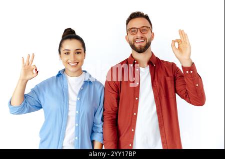 Giovane uomo e donna allegri che gestiscono il segno OK, esprimendo positività e approvazione, vestiti con abiti casual, irradiando fiducia e felicità, in piedi su uno sfondo bianco isolato Foto Stock