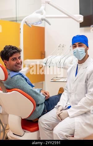 Giovane dentista maschio che indossa una maschera chirurgica e guanti, posa con sicurezza con un paziente sorridente seduto comodamente in un dentista bianco e rosso brillante Foto Stock