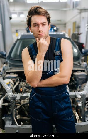 Ritratto verticale del meccanico pensivo maschio in tuta guardare seriamente la fotocamera non sapere quale riparazione durante la manutenzione dell'auto in officina. Foto Stock
