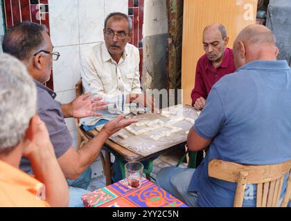 Gruppo di anziani che giocano a domino in un rustico cafй all'aperto ad Alessandria, Egitto. Una scena di strada calda e sociale che cattura la vita quotidiana locale. Foto Stock