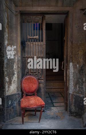 Una sedia in velluto rosso con agenti atmosferici si trova all'esterno di una vecchia porta rustica con cancelli in ferro ad Alessandria, Egitto, che evoca un'estetica urbana vintage. Foto Stock