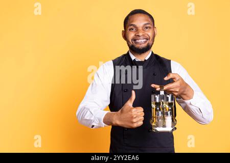 Cameriere cordiale che dà il simbolo del pollice davanti alla telecamera, presentando il suo accordo e le emozioni positive in studio. Il personale giovane dà un simile e porta il cruet set per i tavoli. Foto Stock