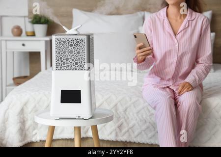 Moderno umidificatore d'aria sul tavolo contro la donna che usa il telefono cellulare in camera da letto, primo piano Foto Stock