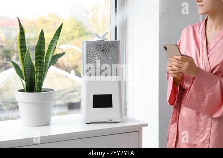 Donna che utilizza un telefono cellulare vicino all'umidificatore d'aria in modalità comoda a casa Foto Stock