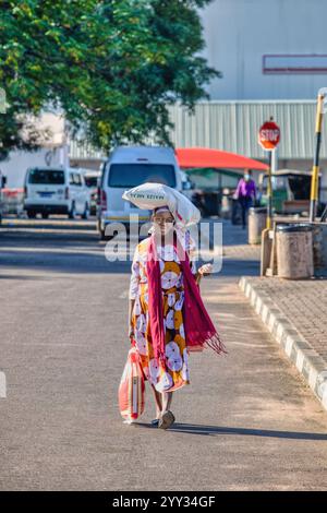 una donna africana porta un sacco di mais sulla testa in strada, sorriso felice sul viso, spesa Foto Stock