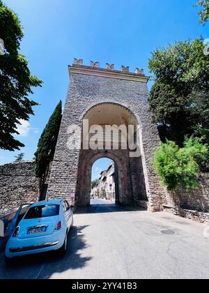 Assisi, Italia - 5 luglio 2022: L'arco in pietra invita i visitatori in un affascinante villaggio europeo, con vegetazione lussureggiante e un'auto parcheggiata sotto il cielo blu Foto Stock