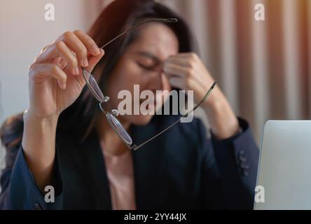 Giovane donna asiatica che sente l'emicrania alla testa. Donna d'affari stanca e sovraccarica di lavoro mentre lavorava al computer portatile in ufficio. Messa a fuoco selettiva Foto Stock