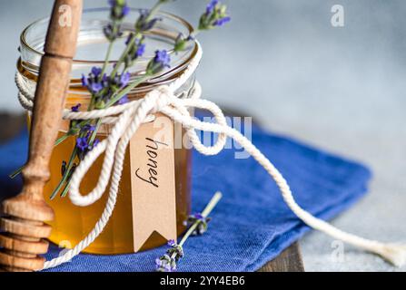 Un vaso di vetro di miele dorato adornato con un fiocco di spago, piccoli rametti di lavanda e un'etichetta scritta a mano, Un cucchiaio di miele di legno poggia accanto ad esso su un panno blu Foto Stock