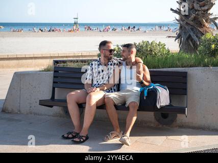 Giovani coppie gay che si godono una giornata di sole in spiaggia, seduti su una panchina indossando abiti estivi casual, abbracciano amorevolmente, mostrando un momento di pubbliche relazioni Foto Stock