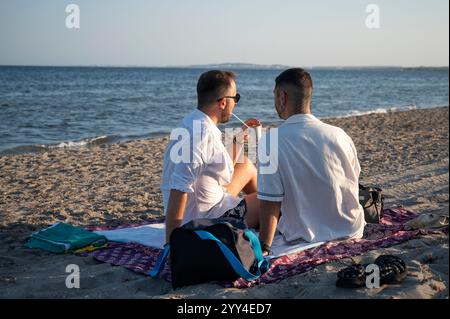 Giovani coppie gay si rilassano sulla spiaggia, condividendo un drink e godendosi un tramonto tranquillo, simboleggiando l'amore e la solidarietà in un ambiente tranquillo sulla costa Foto Stock