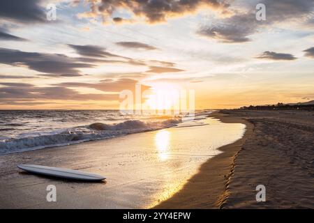 Una tavola da surf poggia su una spiaggia sabbiosa mentre il sole tramonta, che proietta una calda tonalità dorata sulle onde e sul cielo. Il tranquillo paesaggio marino evoca un senso di pace e Foto Stock