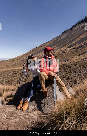 Una coppia di escursionisti che fanno una pausa su un sentiero montuoso sono seduti su una roccia che regge pali da trekking, godendosi l'ampia vista sotto lo skie limpido Foto Stock