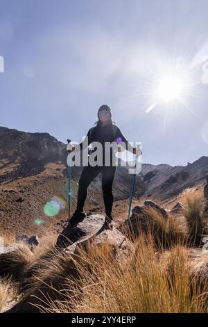 Un escursionista si erge con sicurezza su un affioramento roccioso, tenendo in mano i pali da trekking il sole splende brillantemente, gettando le lenti brillanti, illuminando l'aspra montagna Foto Stock