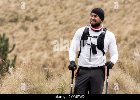 Un uomo di colore, dotato di attrezzatura per arrampicata, sorride tenendo in mano pali da trekking in un paesaggio aspro e erboso, è vestito con wea atletica Foto Stock