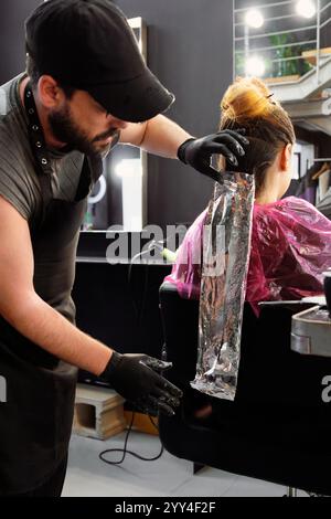Un parrucchiere che indossa i guanti applica il colore dei capelli usando le lamine in un moderno salone il cliente siede su una sedia con un mantello, circondato da un elegante ed elegante de Foto Stock