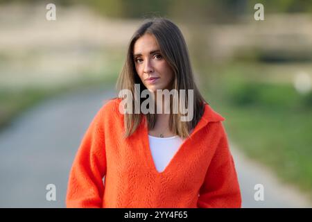 Una donna con un maglione arancione brillante si trova all'aperto con sicurezza, con uno sfondo sfocato del parco sotto l'illuminazione naturale, a mostrare un ambiente informale e sereno Foto Stock