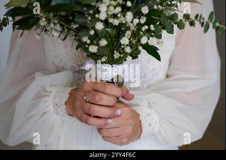 Primo piano di una sposa che regge un bouquet con foglie verdi lussureggianti e delicati fiori bianchi i gambi avvolti in pizzo e un piccolo fascino aggiungono un tocco elegante Foto Stock