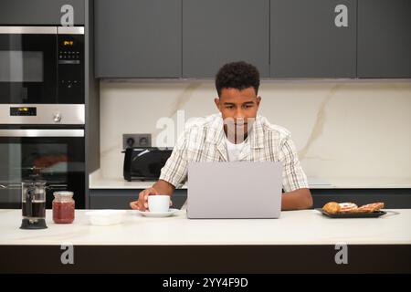 Giovane afroamericano che utilizza un computer portatile in una cucina elegante e moderna bevendo caffè a casa, con cibo al bancone, l'atmosfera è accogliente e rilassante Foto Stock