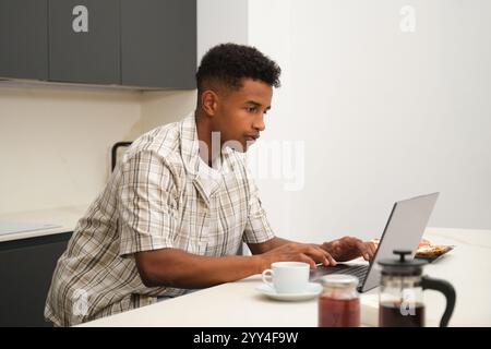 Un giovane afroamericano gode di momenti tranquilli a casa, mentre l'uomo si concentra su un computer portatile, mentre la tranquilla e moderna cucina aggiunge un tocco di intimità Foto Stock