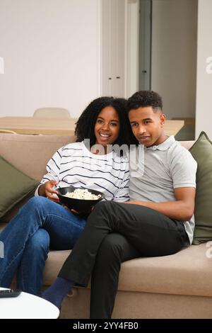 Una giovane coppia di neri si rilassa su un accogliente divano, condividendo una ciotola di popcorn e sorridendo calorosamente, l'ambiente sereno cattura un momento di amicizia e di comunicazione Foto Stock